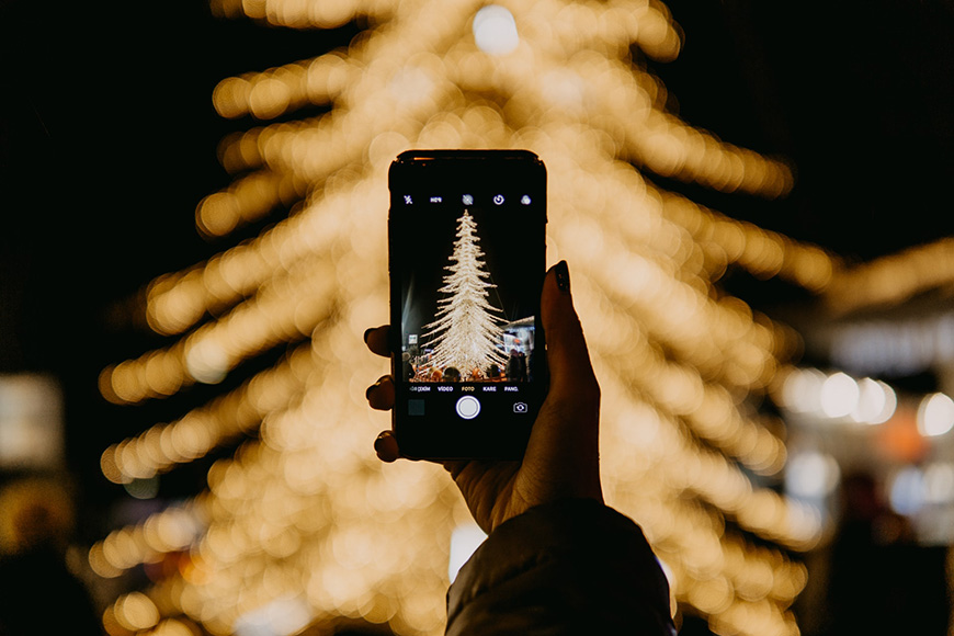 A women holding a smart phone and taking a photo of a Christmas Tree covered in lights