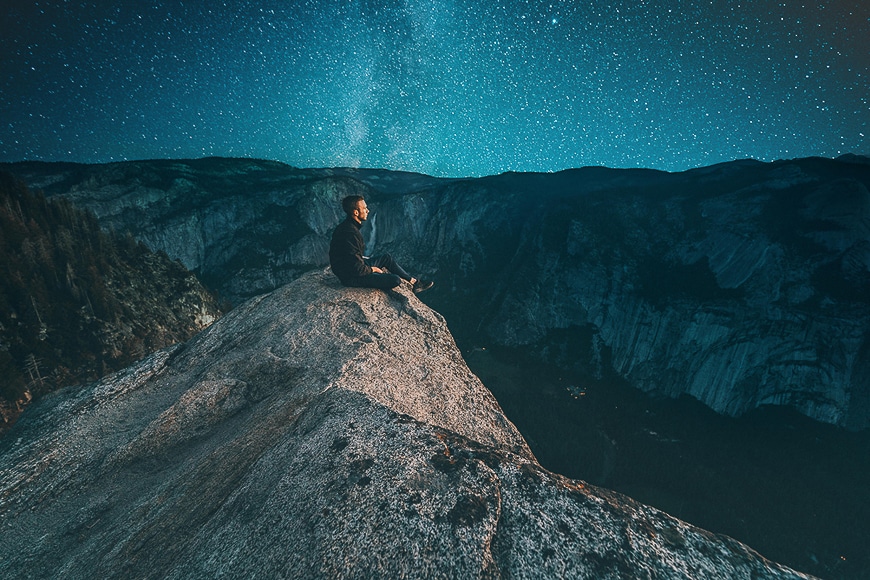 Man sitting on a cliff top with a glowing deep sky backdrop
