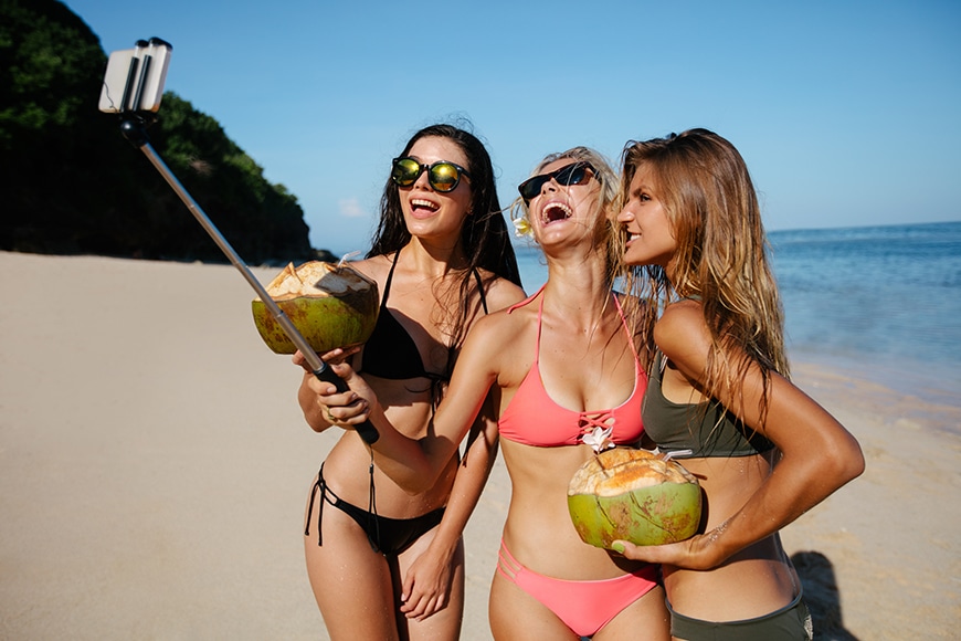 Group of happy young woman with coconuts taking selfie on the beach. Three young women in bikini on the sea shire taking self portrait with smart phone.