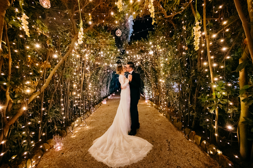 A bride and groom kissing under a string of lights in a garden.