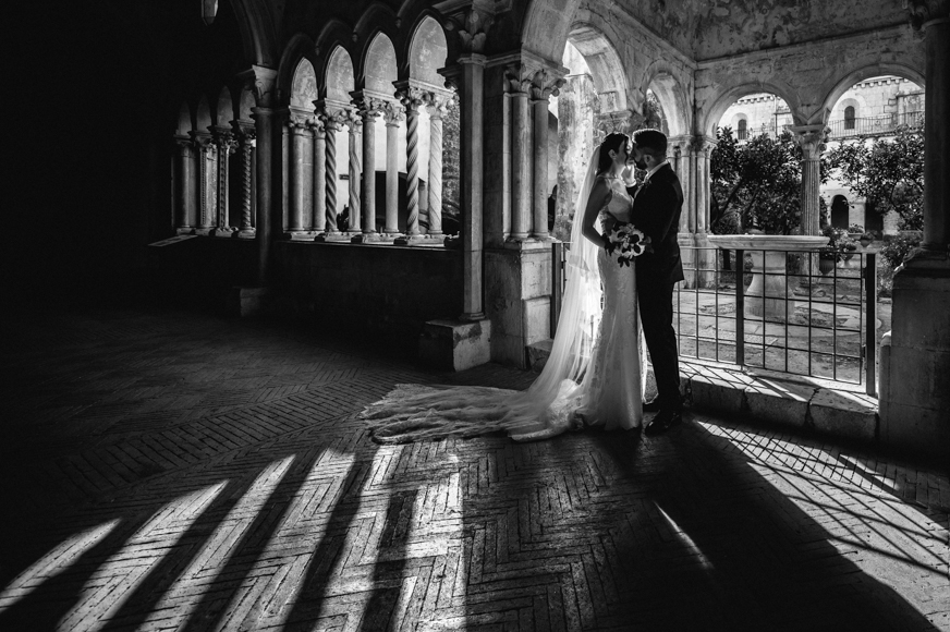 A bride and groom are standing in the shadows of a church.