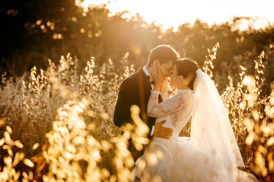 A bride and groom kissing in tall grass at sunset.