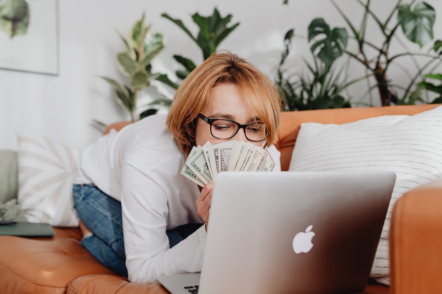 a woman laying on a couch with a laptop and money in front of her.