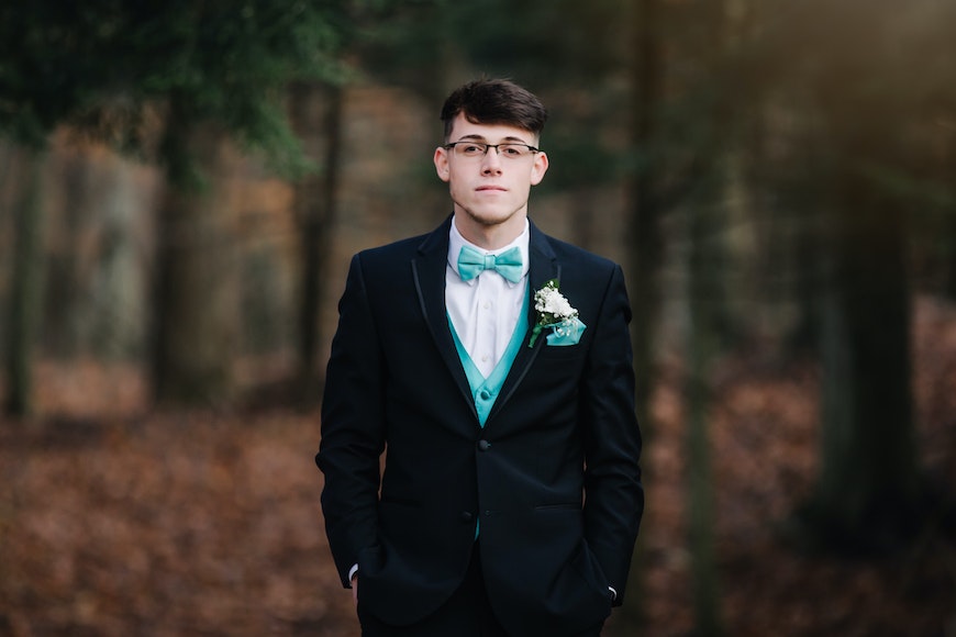 a young man in a tuxedo standing in a forest.