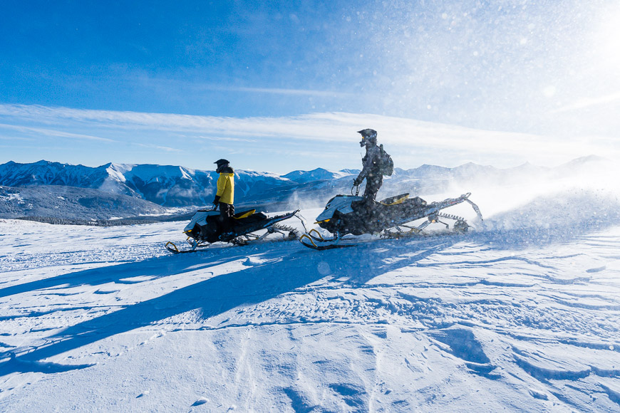 two people on snowmobiles in the snow with mountains in the background.