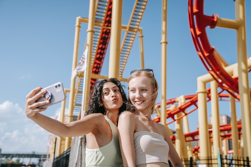 two women taking a selfie in front of a roller coaster.