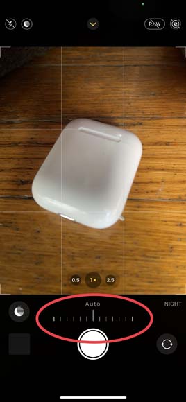screenshot of the iPhone camera app showing an airpods case on a wooden table with a red circle around the exposure settings. 