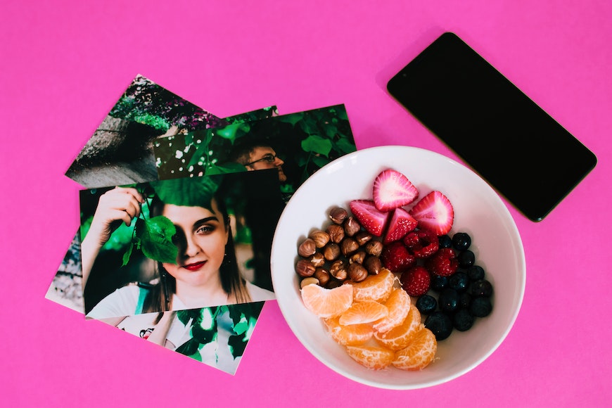 A bowl of fruit, a cell phone, and some photos on a pink background.