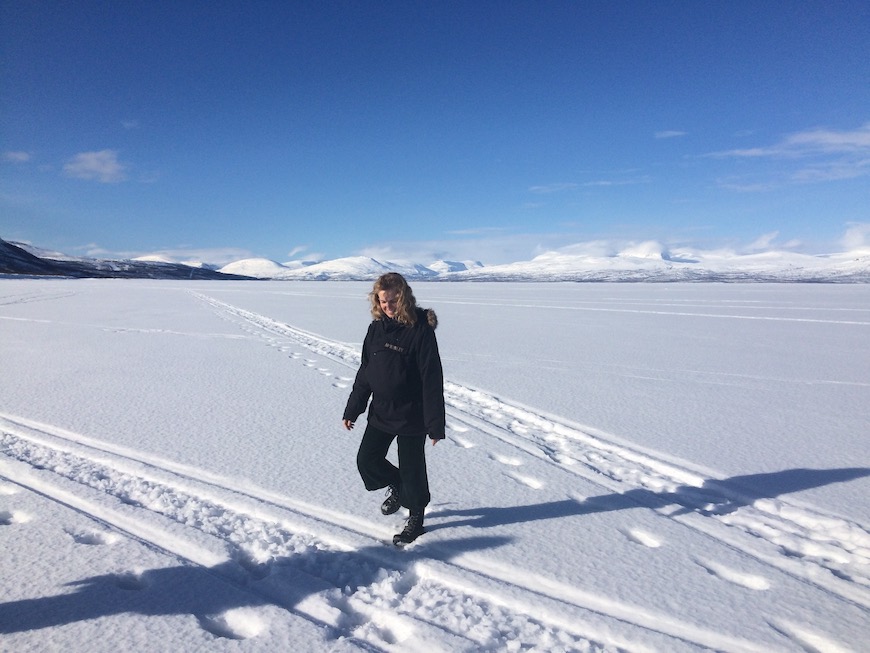 A woman walking across a snow covered field with mountains in the background.