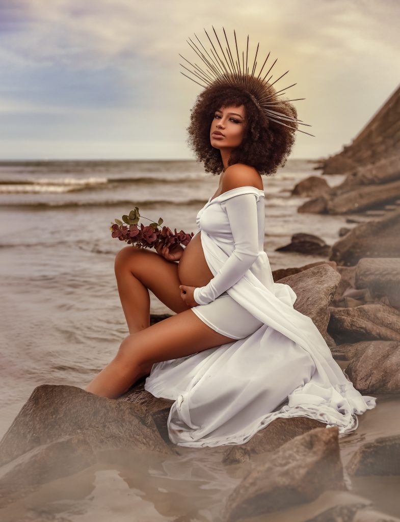 a pregnant woman in a white dress sitting on rocks at the beach.