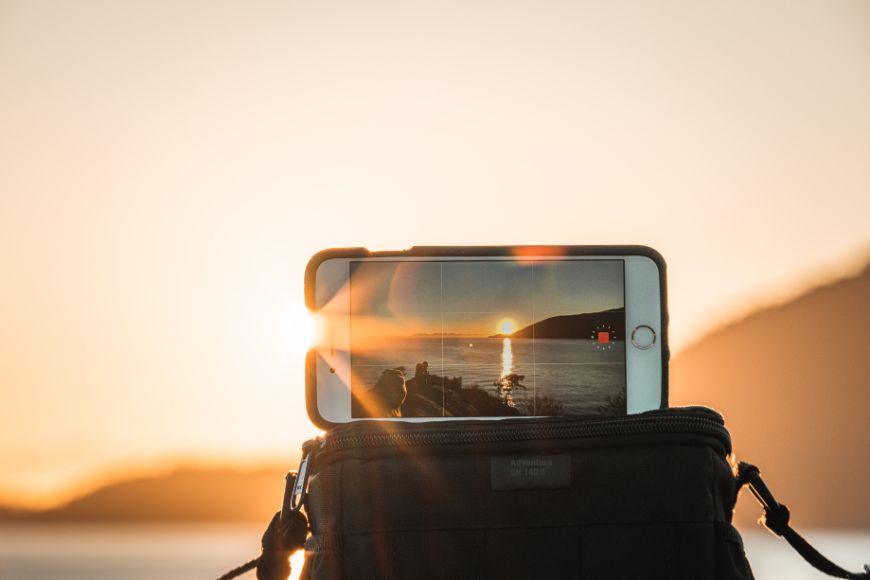 A phone sitting on top of a backpack with the sun setting behind it.