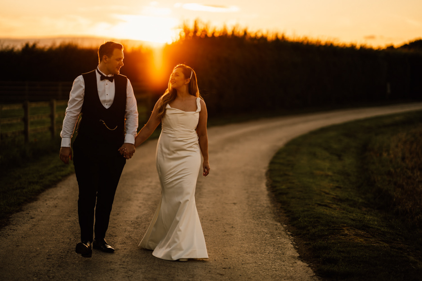A bride and groom walking down a country road at sunset.