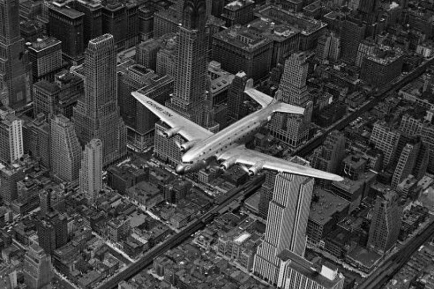 A black and white photo of an airplane flying over a city.