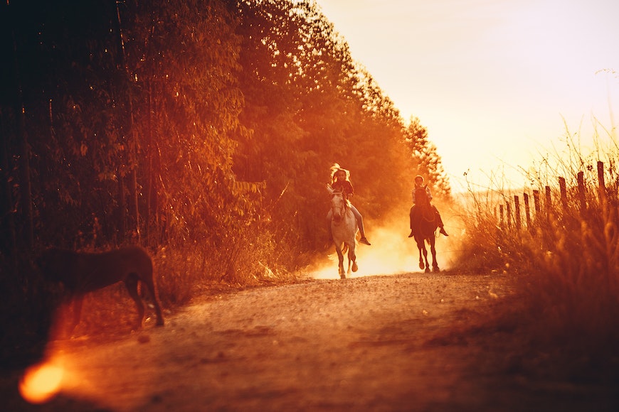 Two people riding horses down a dirt road at sunset.