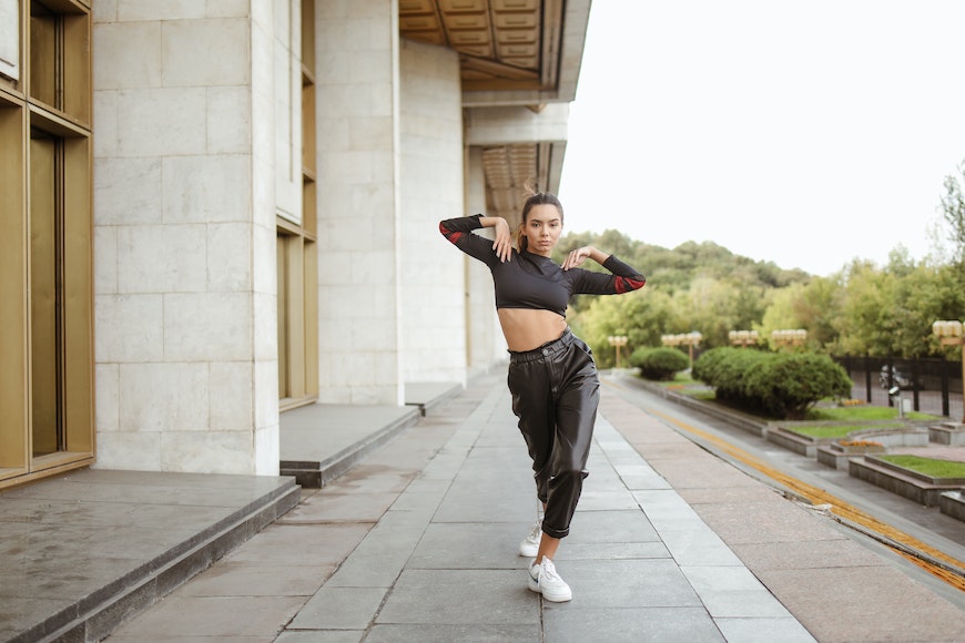 A woman in a black crop top and leather pants is posing in front of a building.