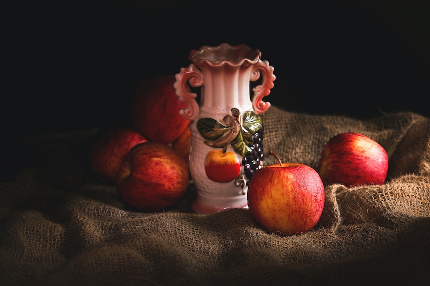 A pink vase sits next to a bunch of red apples.