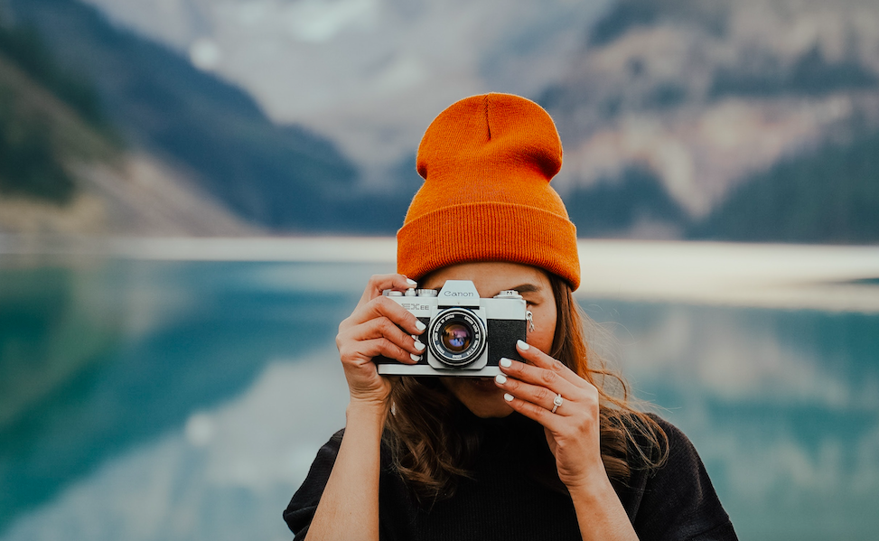 A woman wearing an orange beanie taking a picture of a lake.
