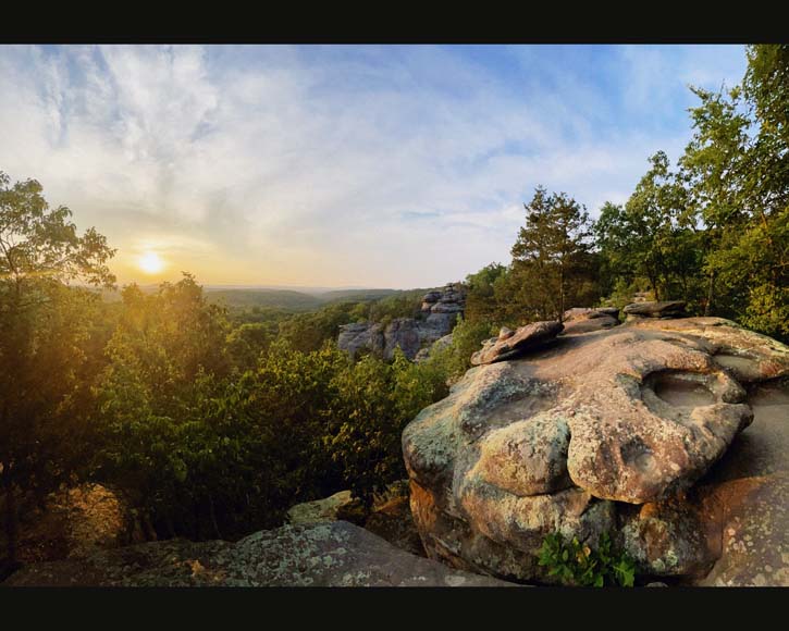 The sun is setting over a rock formation in a wooded area.