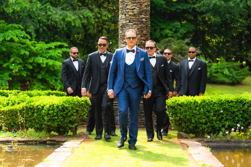 A group of groomsmen in tuxedos walking through a garden.