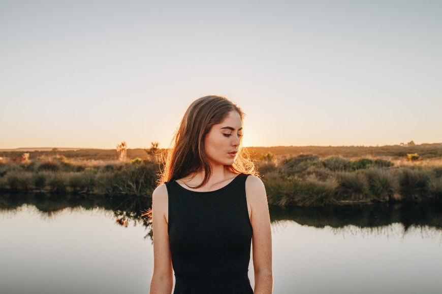 A young woman in a black dress standing in front of a river at sunset.
