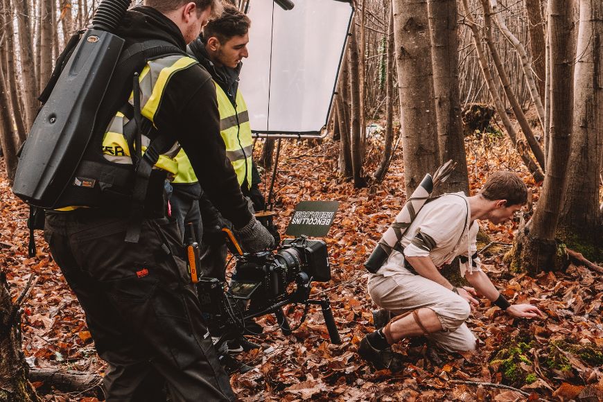 A man is kneeling down in the woods with a camera.