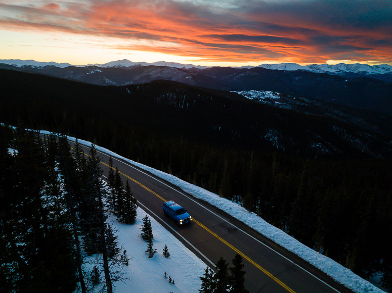 A blue truck driving down a snowy road at sunset.