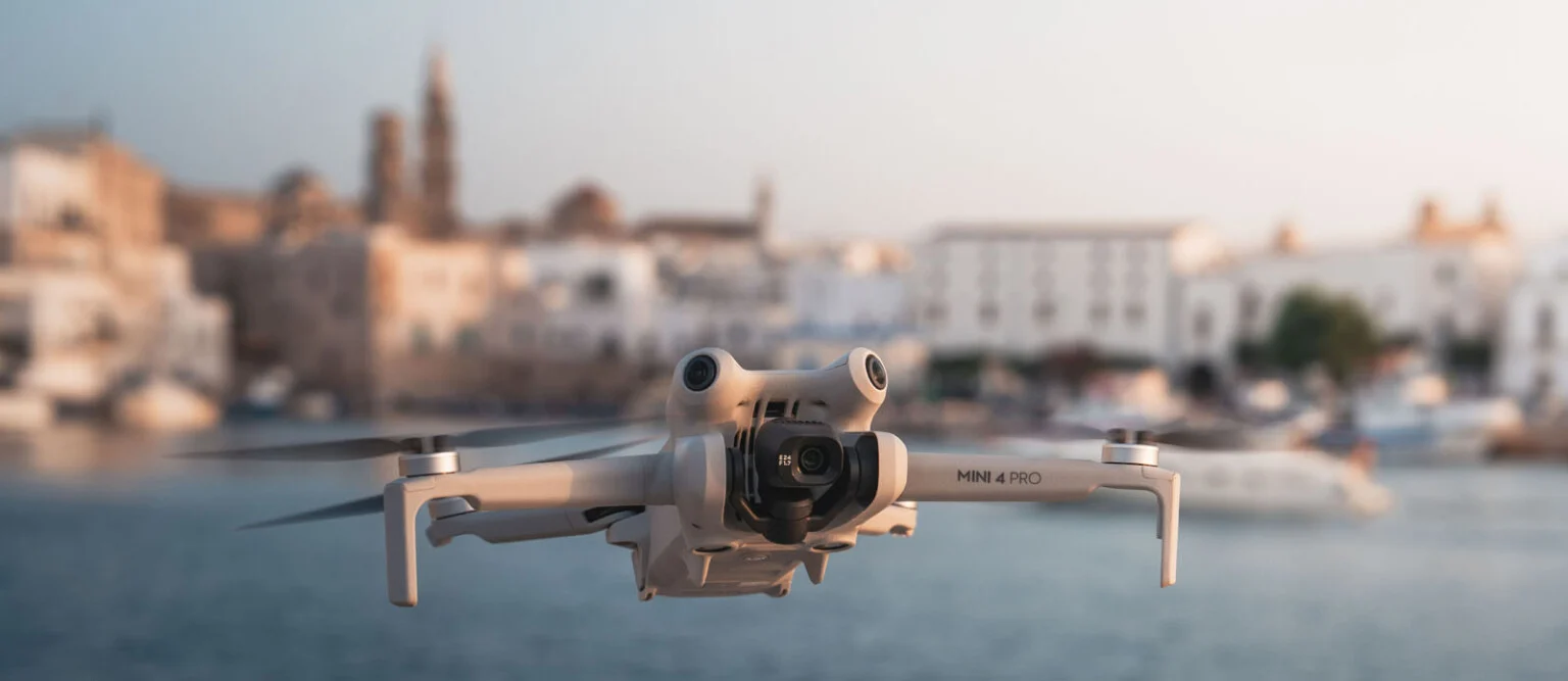 A white drone flying over a city with buildings in the background.