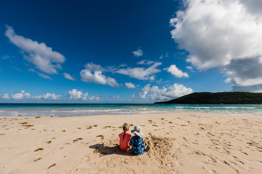 Two people sitting on the sand on a beach.