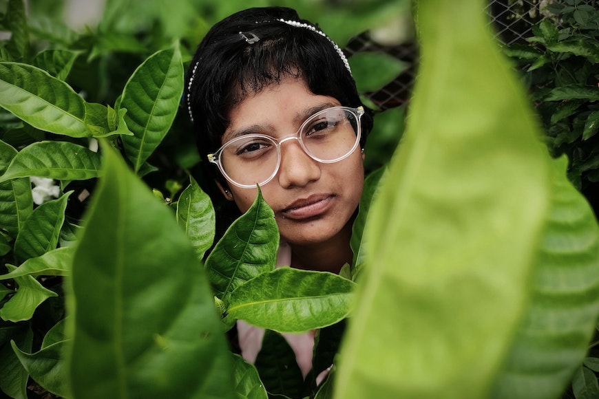 A girl with glasses peeking out of a green leaf.