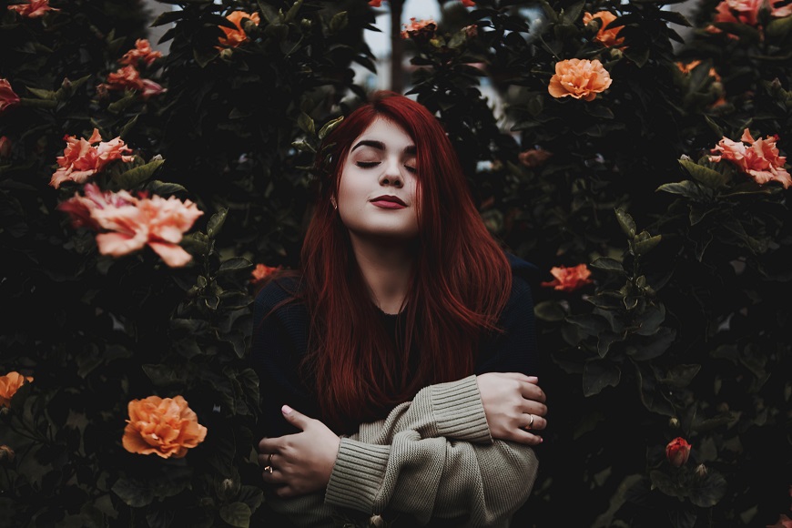 A woman with red hair posing in front of flowers.
