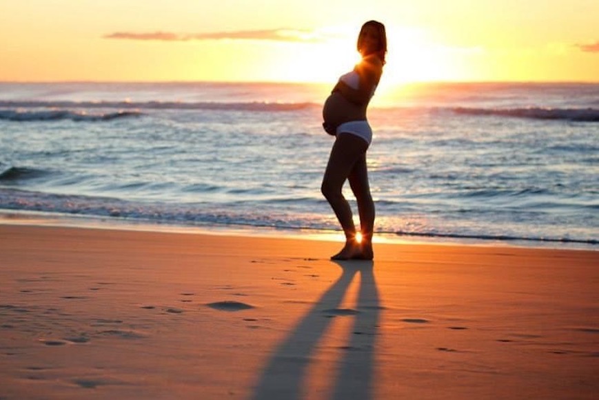 A pregnant woman standing on the beach at sunset.