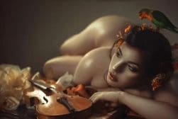A nude woman laying on a violin with a parrot on her head.