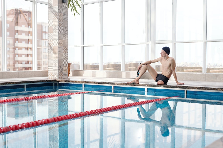 A man is sitting on the edge of an indoor swimming pool.