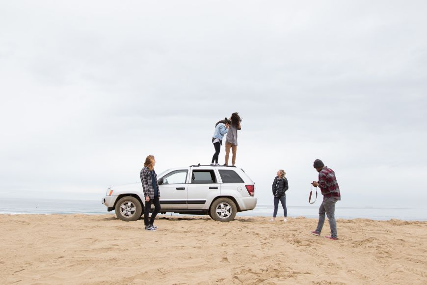 A group of people standing on top of a suv on the beach.