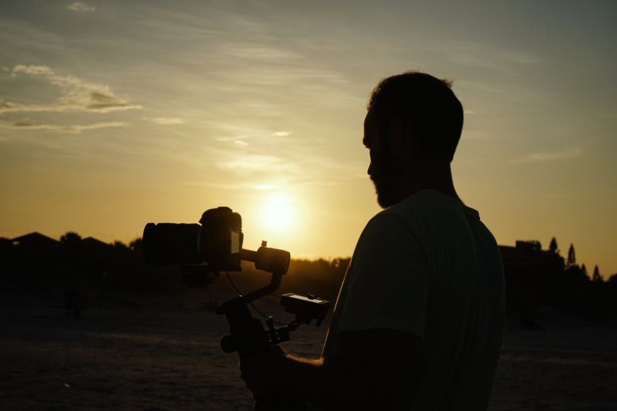 A man holding a camera on the beach at sunset.
