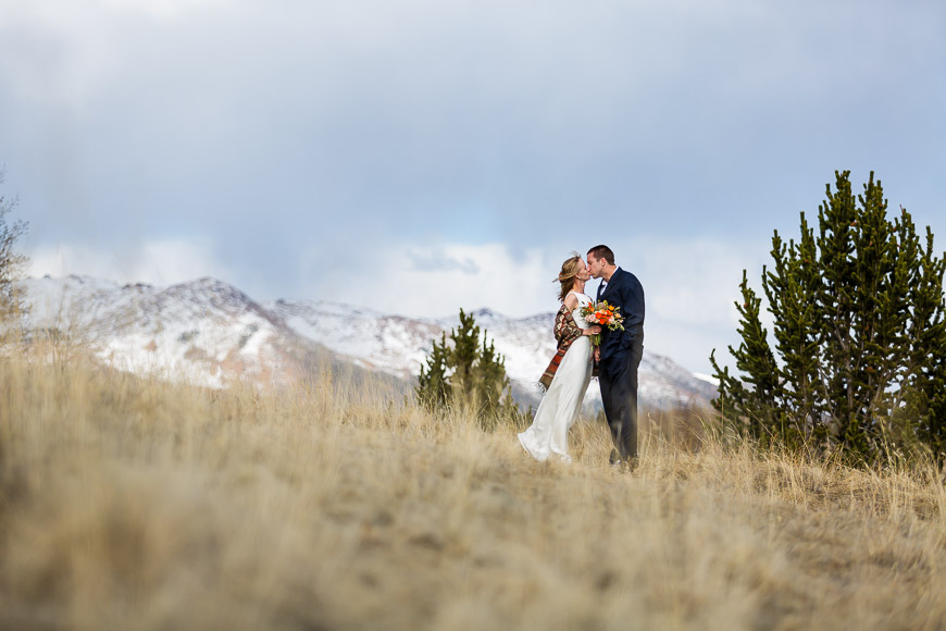 A bride and groom kissing on a grassy field with mountains in the background.