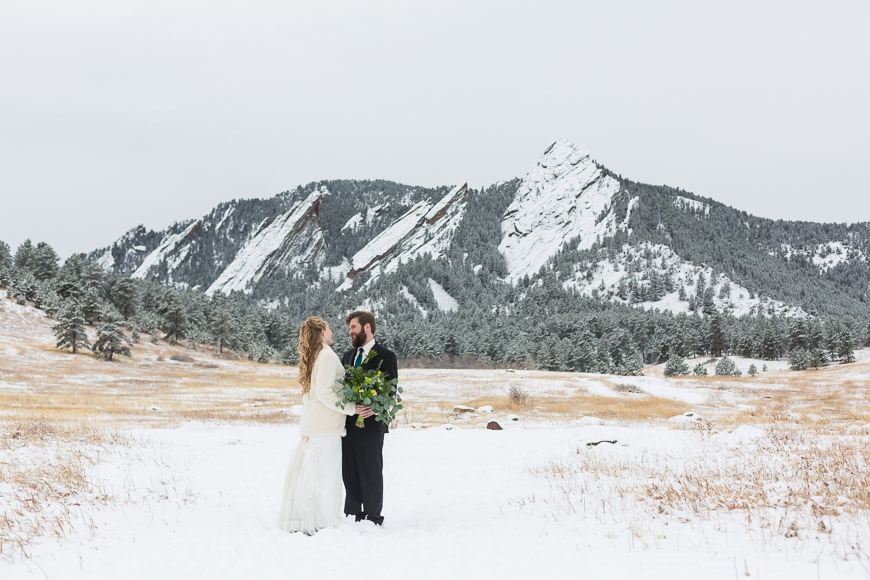 A bride and groom standing in a snowy field with mountains in the background.