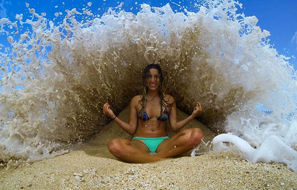 A woman in a bikini sitting on the beach in front of a wave.