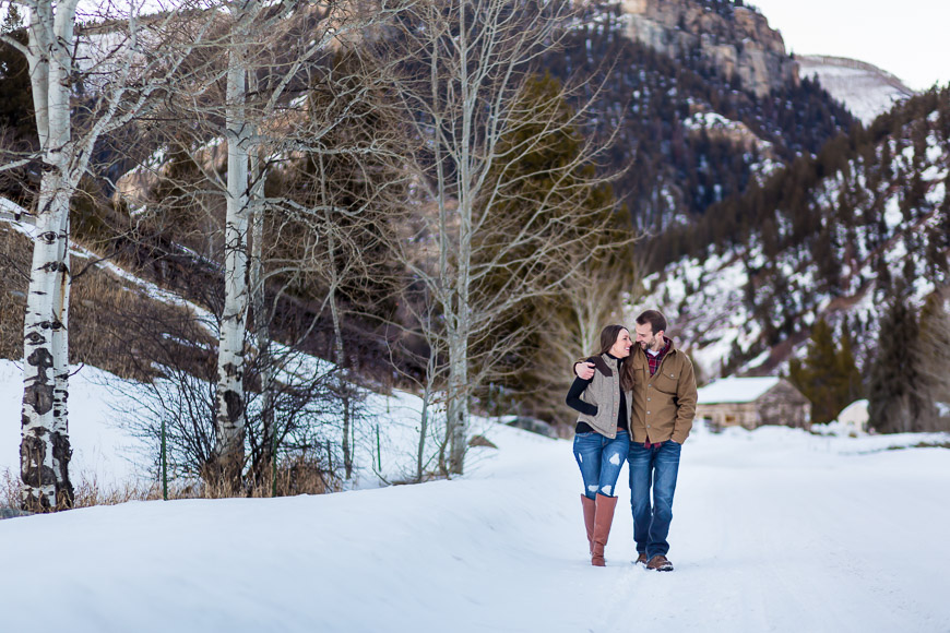 A couple walking down a snowy road with mountains in the background.