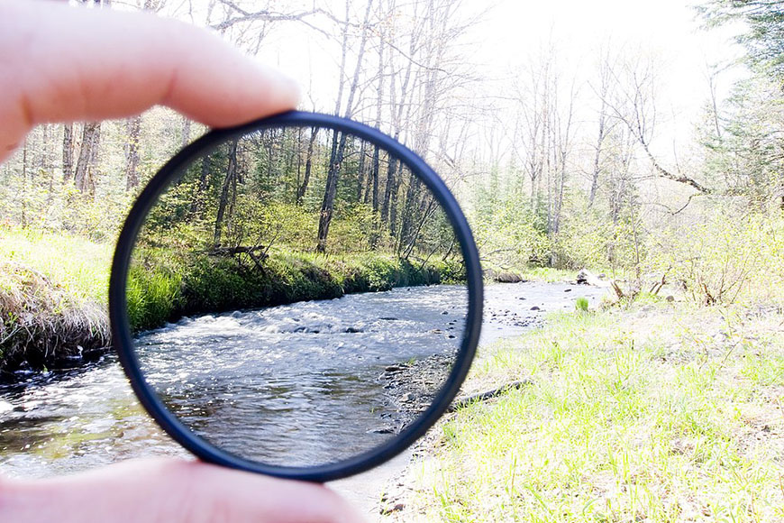 A person holding up a circular lens in front of a river.