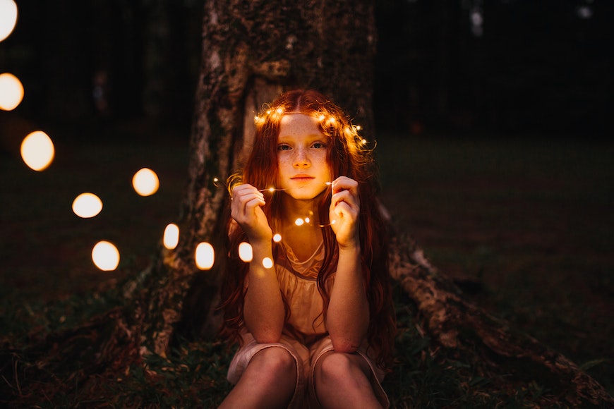 A girl with red hair sitting next to a tree with lights in her hair.