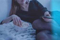 A woman sitting on a bed with a bowl of popcorn and a remote control.