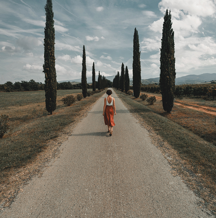 A woman walking down a road with cypress trees.