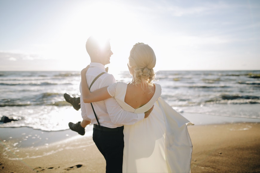 A bride and groom standing on the beach holding hands.