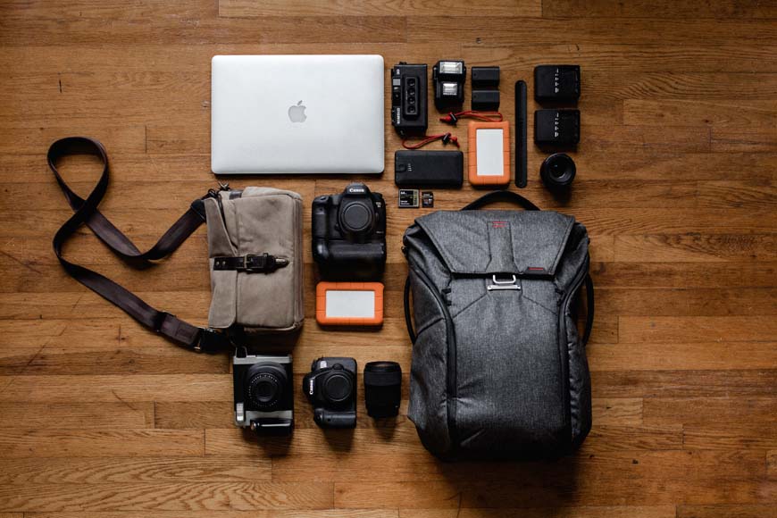A backpack with a laptop, camera, and other items laid out on a wooden floor.