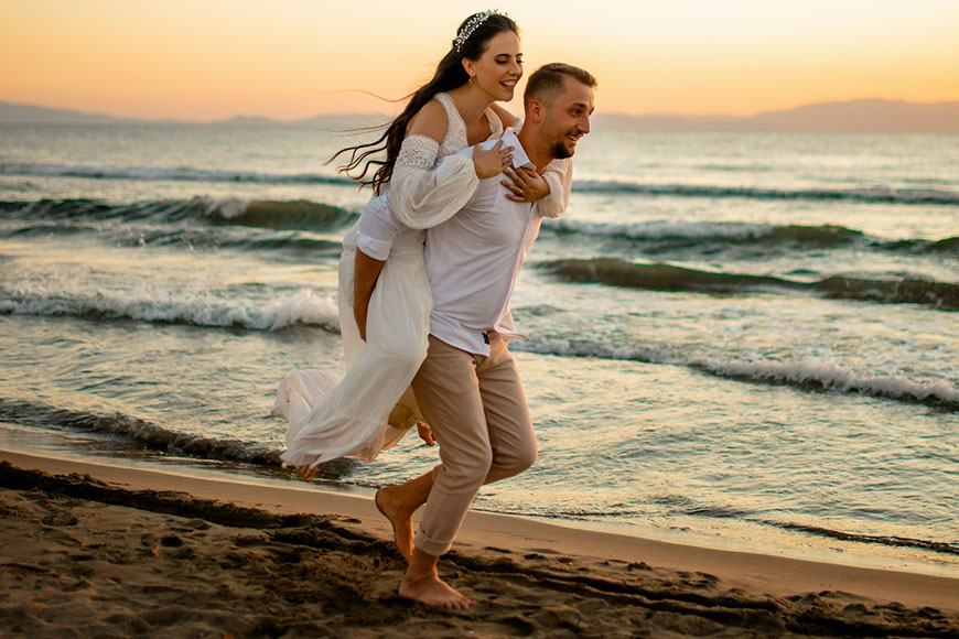A bride and groom carrying each other on the beach at sunset.
