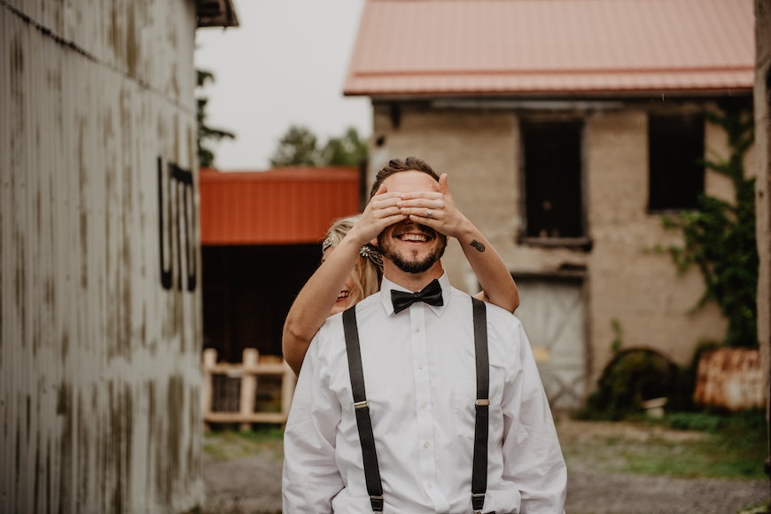 A bride and groom wearing suspenders in front of a barn.
