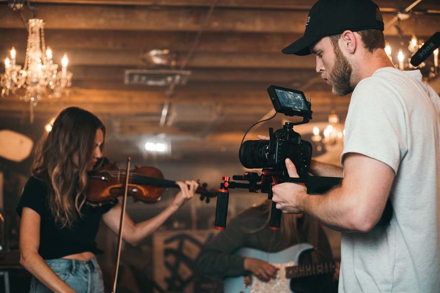 A man holding a camera while a woman plays violin.