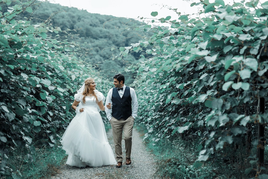 A bride and groom walking down a path in a vineyard.