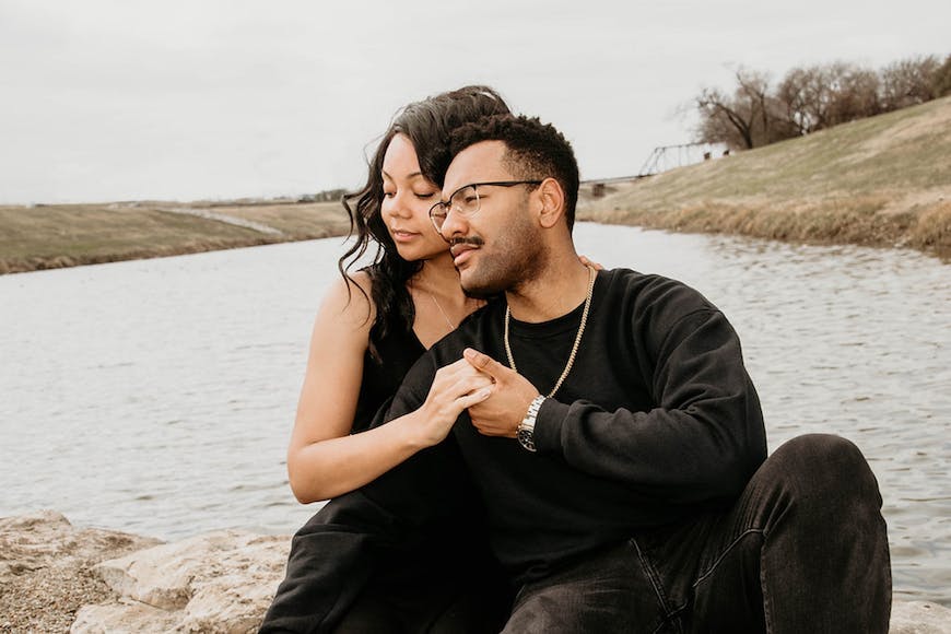 A couple sits on rocks near a body of water during their engagement session.